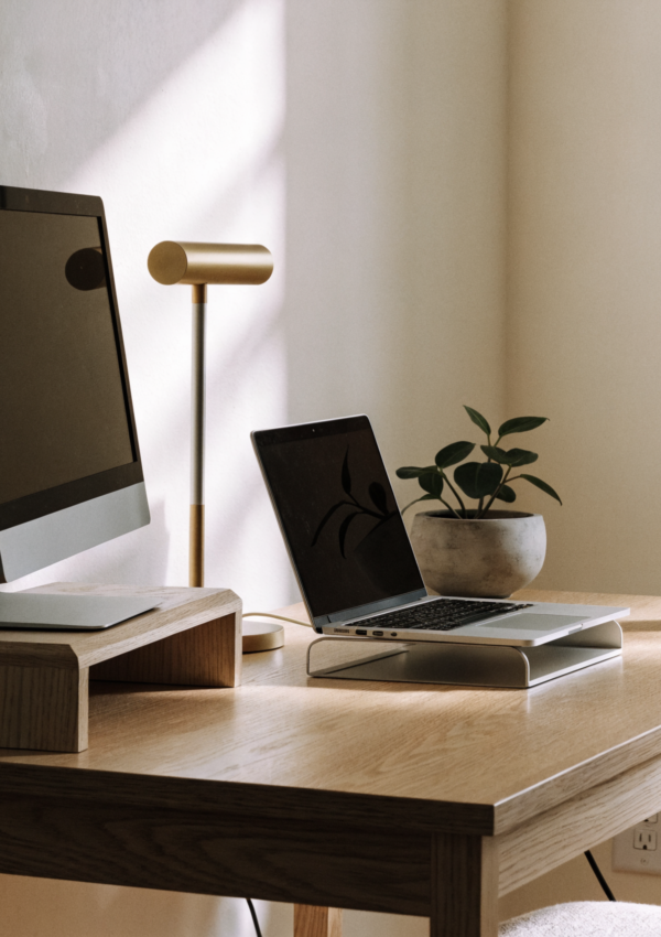 A tidy home office corner with a monitor riser, desk lamp and laptop stand in warm daylight