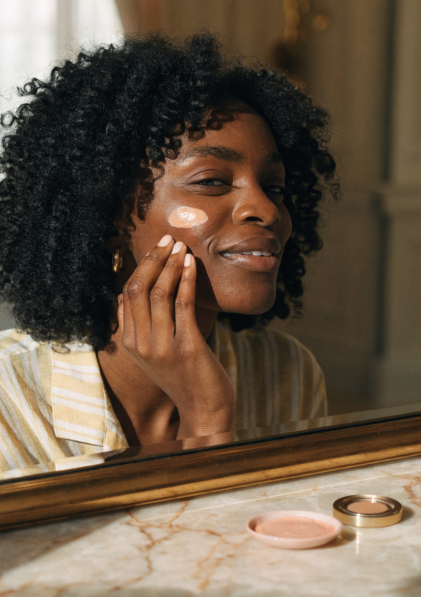A woman with natural curls applying cream blush at a sunlit vanity