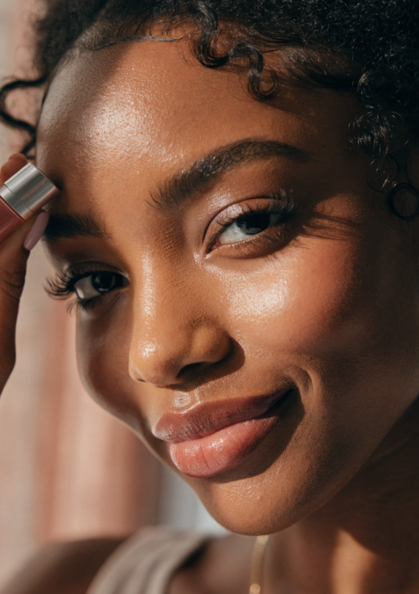 A woman with natural coils holding a tinted lip balm in soft window light