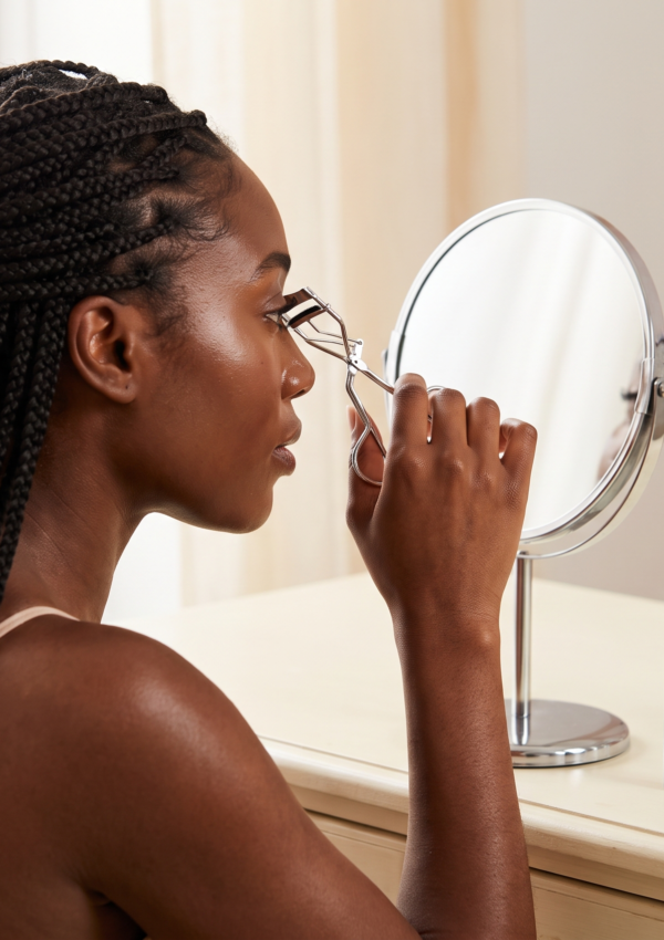 A Black woman with knotless box braids curling her lashes at a cream vanity in warm natural light